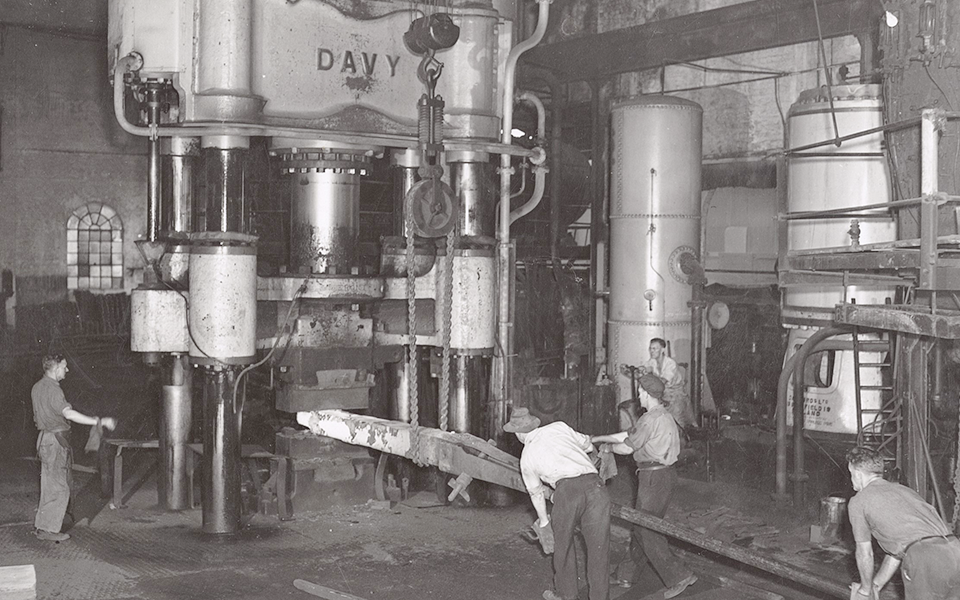 Davy Press in operation. Workers using billet holder to move heated billet under press, crane with pulley and chains attached to hold billet above, operator on left, hydraulic reservoir and steam intensifier visible in right of image. Davy Press in operation. Workers using billet holder to move heated billet under press, crane with pulley and chains attached to hold billet above, operator on left, hydraulic reservoir and steam intensifier visible in right of image.